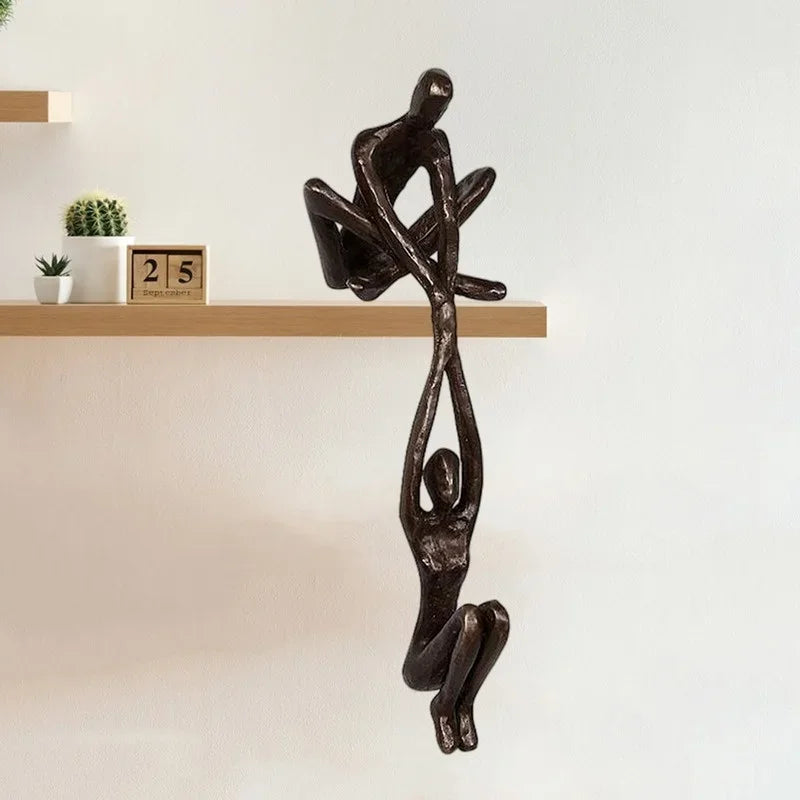 Bronze-tone resin sculpture of two abstract figures balancing together, mounted on the edge of a wooden shelf