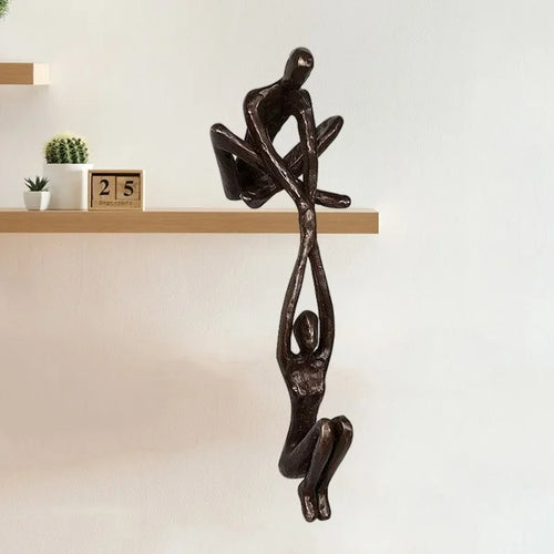 Bronze-tone resin sculpture of two abstract figures balancing together, mounted on the edge of a wooden shelf
