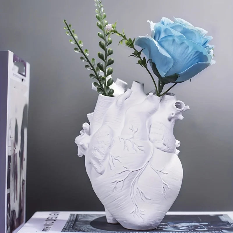 White anatomical heart vase holding a blue rose and greenery beside a book